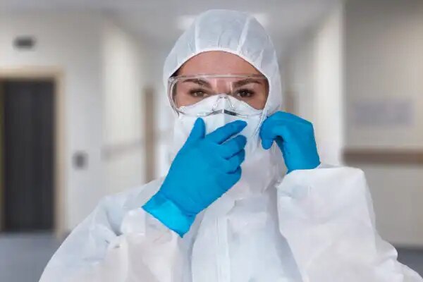 Frau mit Schutzanzug, Schutzbrille, Handschuhe sieht in die Kamera, Klinikgang unscharf im Hintergrund